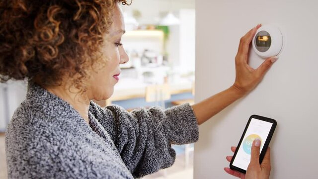 Close up of mature woman wearing dressing gown using app on mobile phone to adjust digital central heating thermostat at home - shot in slow motion