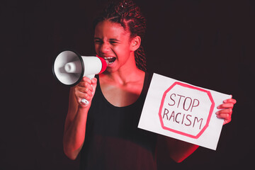Angry African-American girl with poster and megaphone on dark background. Stop racism