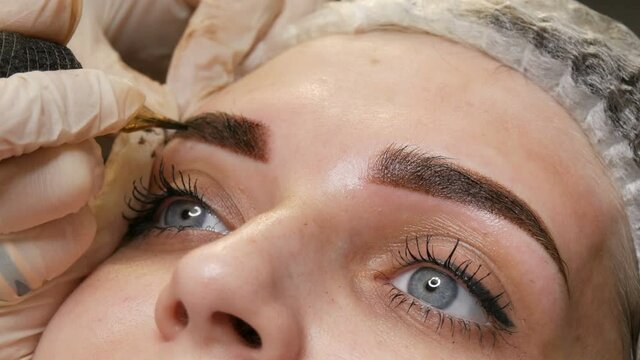 A special needle tattoo machine makes permanent makeup correction of a young woman's eyebrows. A pigment of dark paint is injected under the skin. Microblading, powder spraying close up.