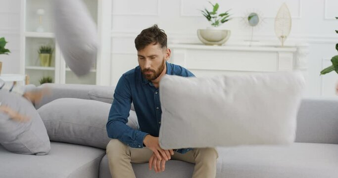 Sad stressed Caucasian handsome man sitting on couch and noisy kids boy and girl running around him and screming. Tired dad with ill-mannered small daughter and son at home.