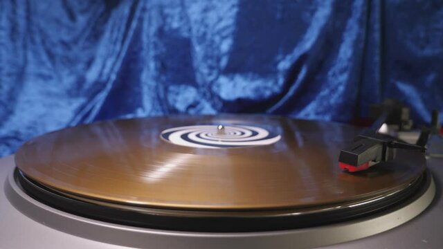 Stylus arm and needle lowered on 12” gold vinyl record on a DJ turntable,  blue velvet background. Retro LP platter. Mid shot.  Hi-fi music equipment for disco, punk, grunge, pop 60s, 70s, 80s, 90s 4K