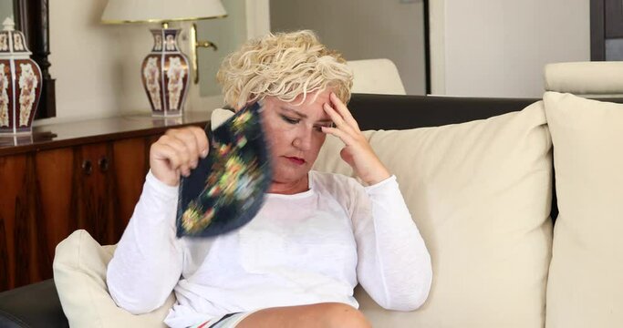 Portrait of a middle aged woman suffering a heat wave using a fan sitting on a couch in the living room at home
