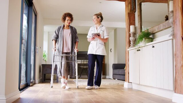 Mature woman in dressing gown using walking frame being helped by female nurse with digital tablet - shot in slow motion
