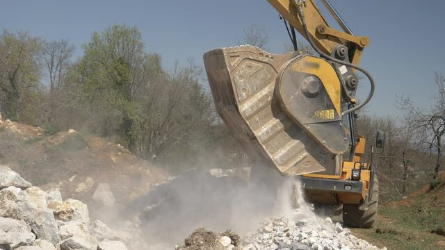 CLOSE UP: Crusher bucket of a yellow excavator demolishes big white rocks into tiny rubble. Industrial machinery demolishing stones into debris at a busy construction site in the sunny countryside.