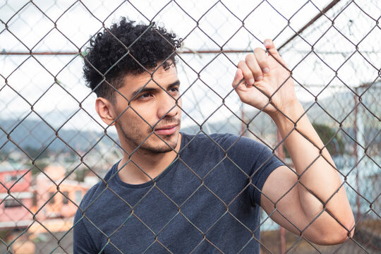 Portrait of young hispanic man in front of a metal mesh at sunset - pensive young man reflecting on the rooftop - urban style
