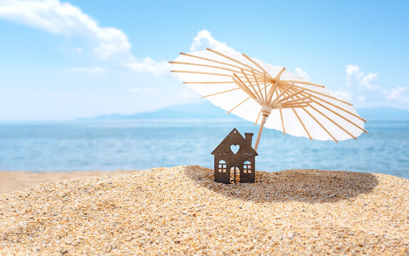 Sun Umbrella And House On Sandy Beach, Blue Sky And Sea In The Background. Investment In Resort Real Estate Concept. Dream Vacation At Sea. Copy Space.