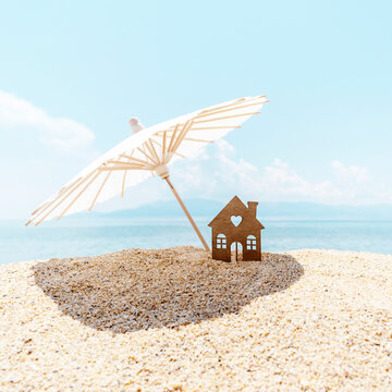 Mminiature house under beach umbrella on seashore on background of blue sky and sea. Concept of dream holiday or real estate on the beach. Square size. Copy space