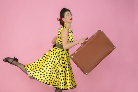 Beautiful pin-up girl on a pink background. Retro woman in yellow polka dot dress. Girl with an old suitcase. Travel concept.