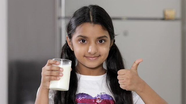 A medium shot of a cute little girl happily showing thumbs up to a glass of milk. A lovable young kid gives a thumbs up looking at the camera while holding a glassful of milk  - lifestyle kids concept