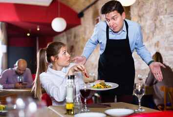 Displeased unhappy aggressive young woman conflicting with apologetic waiter because of poor...
