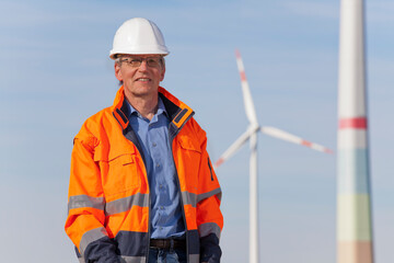 Smiling engineer with hard hat and protective clothing in front of windmills
