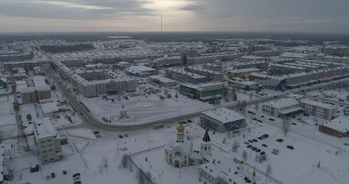 Aerial view of winter city. In the shot are residential buildings and city administration building with square and church. There is snow on the roads, cars drive on the roads. Beyond the city endless 