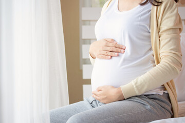 Young beautiful pregnant woman sitting on bed and touching belly near the window in bedroom. Pregnancy, motherhood, people and expectation concept.