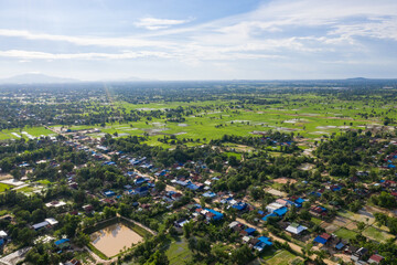 A top down aerial view of a small country town with traditional houses with orange roofs, a red dirt road, rice fields, and palm trees in the jungle in Cambodia.