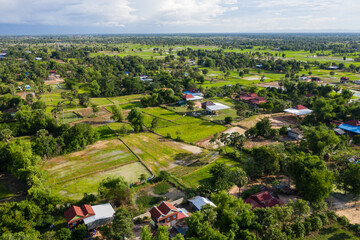 Fototapeta premium A top down aerial view of a small country town with traditional houses with orange roofs, a red dirt road, rice fields, and palm trees in the jungle in Cambodia.