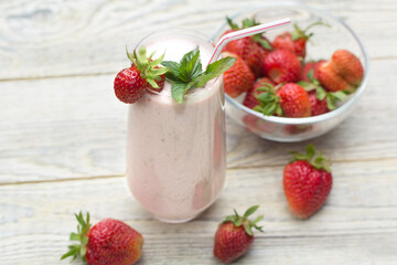 A glass glass with a strawberry milkshake with a straw, stands on a light wooden background, the berries are in a transparent vase and on the table