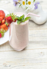 A glass glass with a strawberry milkshake and a white plate with strawberries stand on a light wooden background