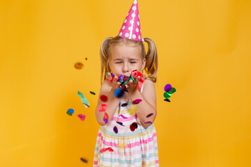 Happy birthday child girl in pink cup blowing confetti on colored yellow background. Celebration, childhood.