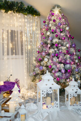 Pine tree decorated with purple bulbes, shiny star and warm garland lights with two white lanterns on the foreground. Merry Christmas, Happy New Year and winter holidays concept.