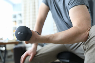Fototapeta na wymiar Close-up of strong man training at home and lifting black dumbbell for muscles. Indoors workout on quarantine. Male in stylish grey shirt. Sport and active lifestyle concept