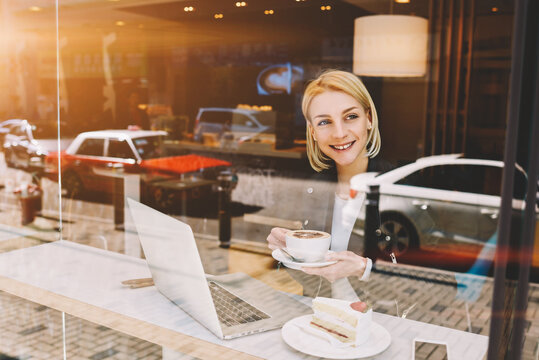 Young entrepreneur is looking satisfied while is thinking about passed business meeting while having work break in coffe shop. Thougfull female freelancer is enjoying cappuccino after work on laptop