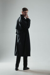 Young and handsome caucasian man posing in a black trench coat in the studio with gray background....