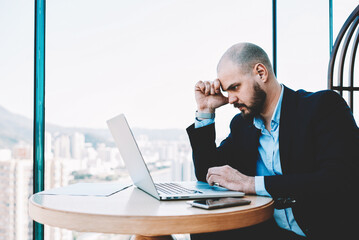 Naklejka premium Serious intelligent banker is spending important banking operation via portable laptop computer. Skilled man architect is working on a new project via net-book, while is sitting in office interior