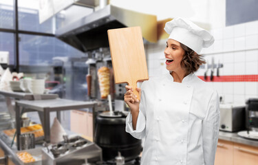 cooking, culinary and people concept - happy smiling female chef in toque with wooden cutting board over restaurant or kebab shop kitchen background