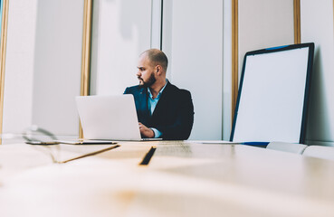 Male lawyer in formal wear creating business strategy during working process at modern laptop device connected to high speed wireess internet sitting in own office interior near bord with mock up area