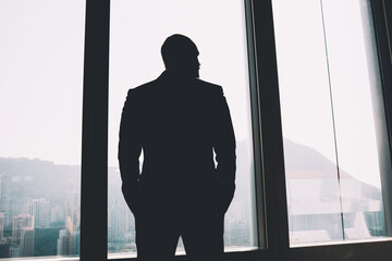 Back view of male banker dressed in black elegant suit holding hands in pockets and looking out of window while thinking on new financial project after hard working day standing in office interior