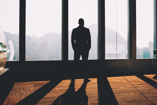 Black silhouette of entrepreneur dressed in formal wear looking out of window and thinking on developing business strategy and new ideas standing in office after hard day and enjoying sunset scenery