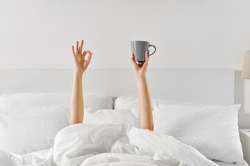 Fototapeta na wymiar morning, comfort and people concept - young woman with cup of coffee lying in bed and showing ok hand sign at home bedroom