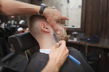 Unrecognizable barber using razor blade, shaving beard of male client
