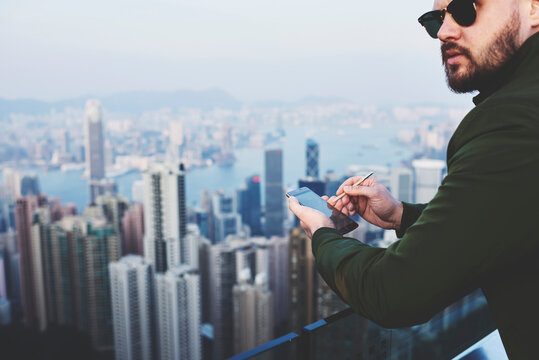 Closely image of a bearded hipster guy in fashionable sunglasses with mobile phone in hand is looking away, while is standing on building roof against blurred business district skyscrapers in New York