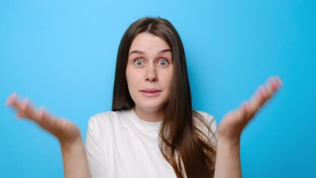 Portrait of upset young girl raising hands in indignant gesture, asking reason of failure, expressing disbelief irritation, wears t-shirt, isolated on blue studio background. Selective focus