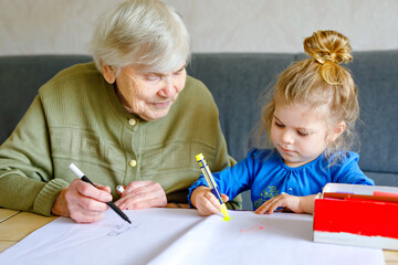 Beautiful toddler girl and grand grandmother drawing together pictures with felt pens at home. Cute child and senior woman having fun together. Happy family indoors - Powered by Adobe