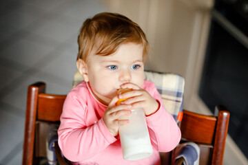 Cute adorable baby girl holding nursing bottle and drinking formula milk. First food for babies. New born child, sitting in chair of domestic kitchen. Healthy babies and bottle-feeding concept