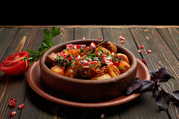 Fried potatoes with meat and pomegranate seeds, in a clay plate, on a wooden background with herbs and tomato
