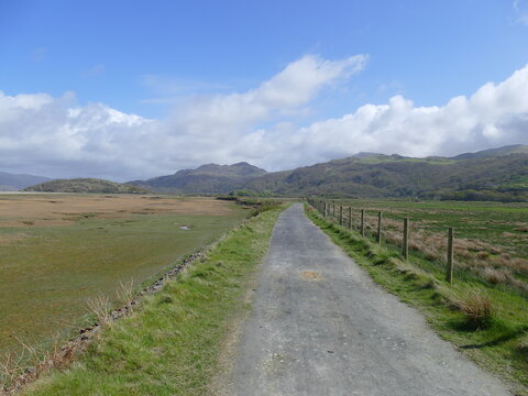 Tarmac footpath crossing a grassy marshy plain with mountains and blue sky in the background near Fairbourne Barmouth Wales