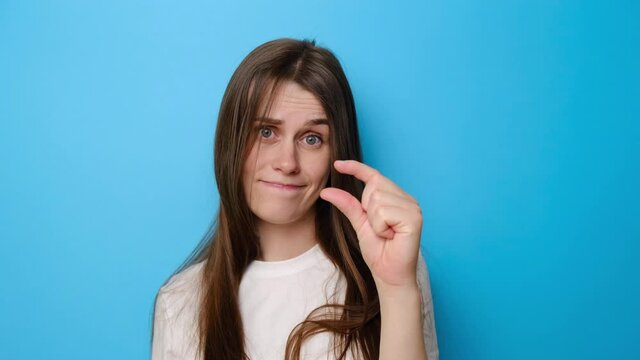 Portrait of brunette woman isolated over blue studio background shows very tiny object, shapes something small, dressed white t-shirt, female looks in puzzlement, need some more. I need little bit