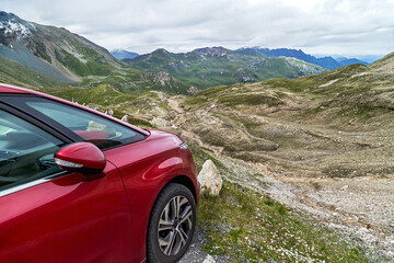Naklejka premium Red car for traveling is standing on the mountain road in the Alps, Austria. Amazing landscape with snowy mountain peaks and sky on background. Family vacation, travel, transportation concept.
