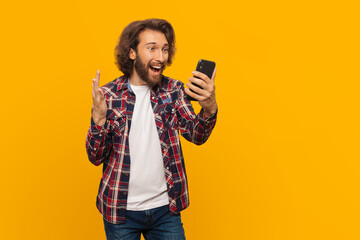 Happy mma man with curly hair in a plaid shirt is very happy about victory looks into a mobile phone and shouts with delight on isolated yellow background