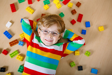 Little blond child with glasses playing with lots of colorful plastic blocks indoor. Kid boy wearing colorful shirt and having fun with building and creating