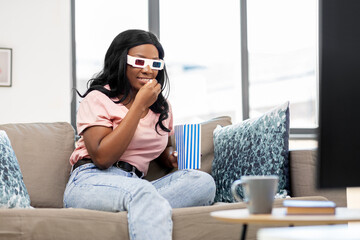 Fototapeta premium people, race, ethnicity and portrait concept - happy smiling african american young woman in 3d glasses watching tv and eating popcorn at home