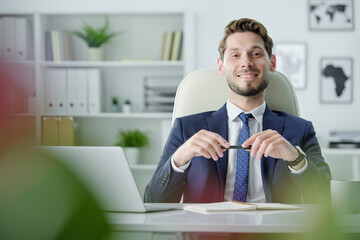 Portrait of smiling confident young businessman with stubble sitting at office table with laptop...