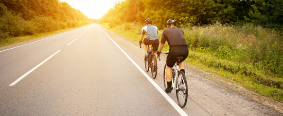 Two cyclist riding through the asphalt road in sunset - obrazy, fototapety, plakaty