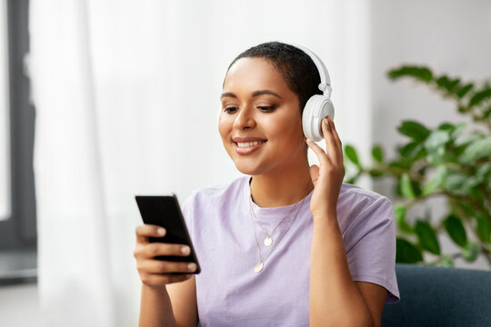 people, technology and leisure concept - happy smiling young african american woman in glasses with smartphone and headphones sitting in chair and listening to music at home