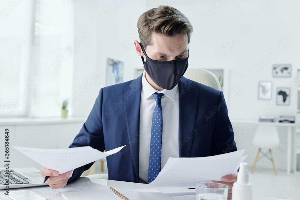 Poster modern young businessman in cloth mask sitting at table in office and reading contract while examini