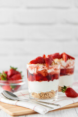 Homemade layered dessert with fresh strawberries, cream cheese or yogurt, granola and strawberry jam in glasses on white wood background. Healthy organic breakfast or snack concept. Selective focus.