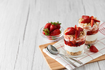 Homemade layered dessert with fresh strawberries, cream cheese or yogurt, granola and strawberry jam in glasses on white wood background. Healthy organic breakfast or snack concept. Selective focus.